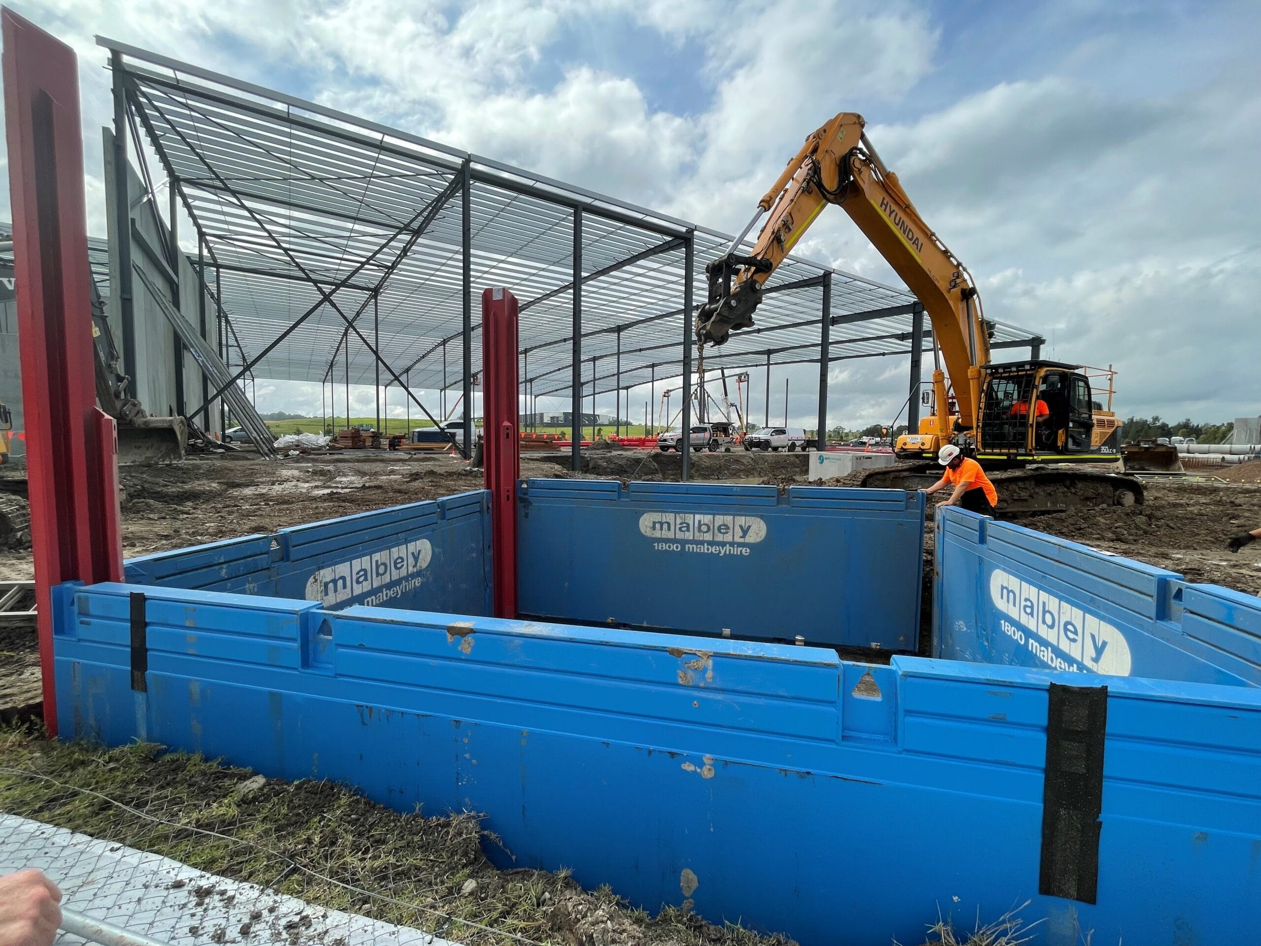 Excavator loading material into Mabey trench shoring shield at construction site